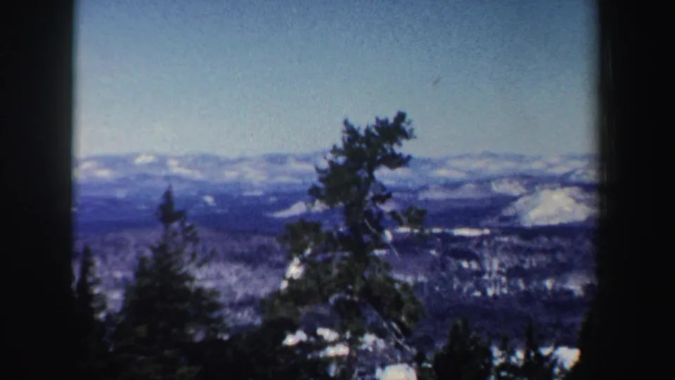 1962 New Hampshire 8mm frame showing evergreen trees, snow patches, distant hills, and a pale winter sky.