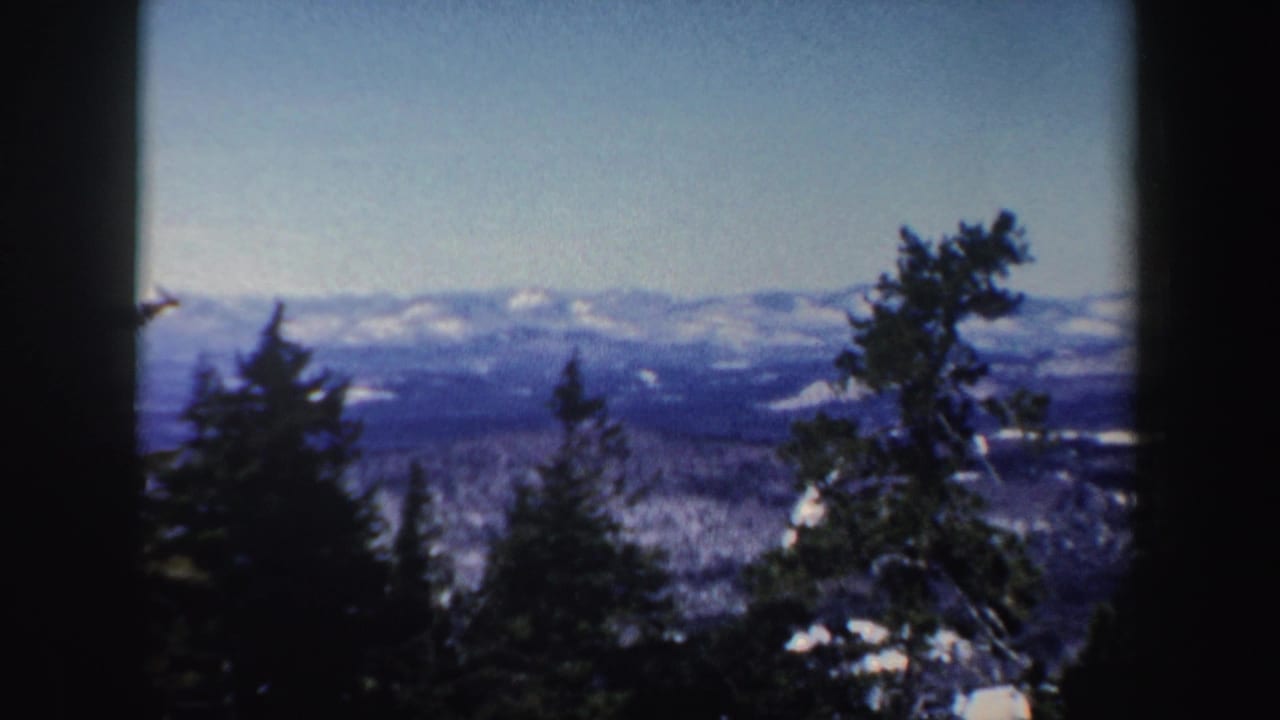 1962 New Hampshire 8mm frame with dark tree trunks in the foreground and a snowy mountain range in the distance.