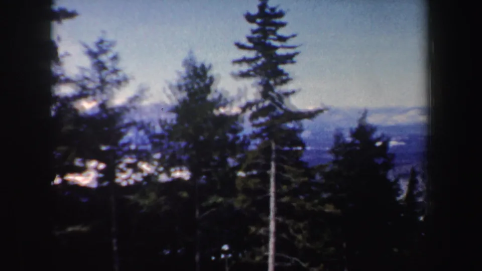 1962 New Hampshire 8mm frame of evergreen trees, bright snow patches, and distant blue-gray winter mountains.