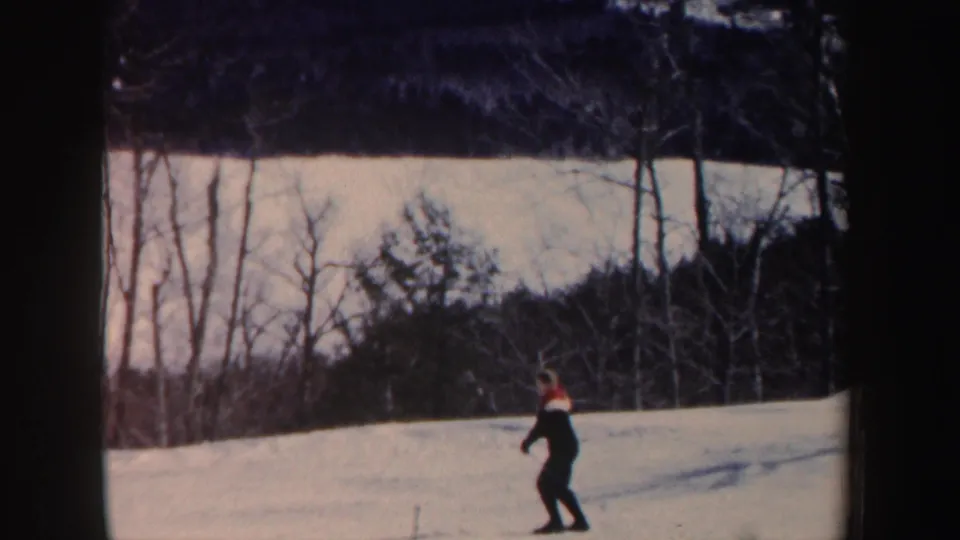 A lone skier in dark clothing appears near the lower part of a snowy New Hampshire slope, with leafless trees and woods behind.