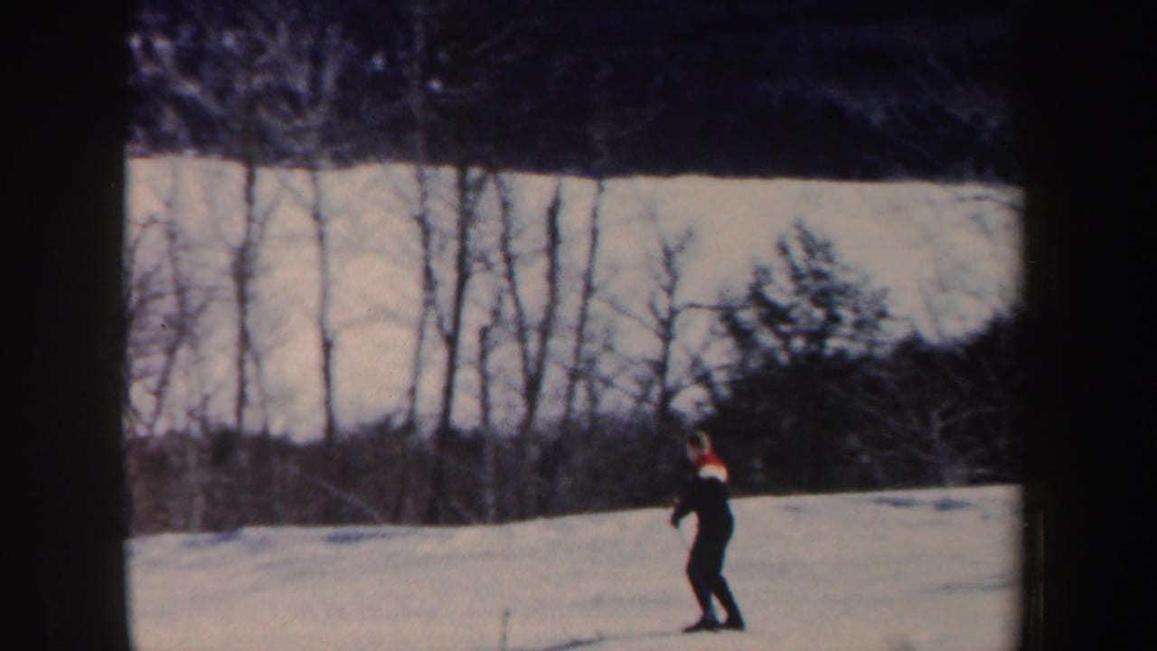 In 1962 New Hampshire home-movie footage, a distant skier in dark clothing moves across a snowy slope backed by bare trees.