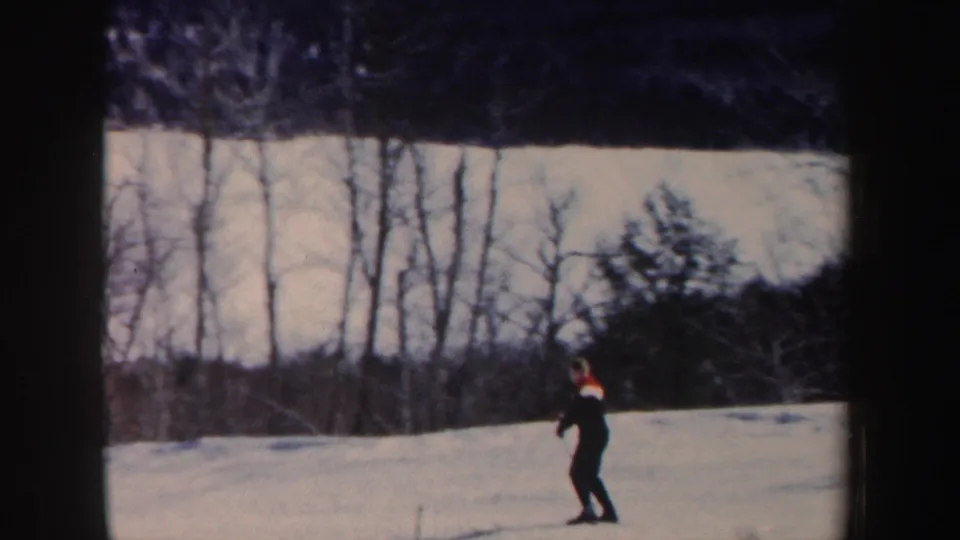 In 1962 New Hampshire home-movie footage, a distant skier in dark clothing moves across a snowy slope backed by bare trees.