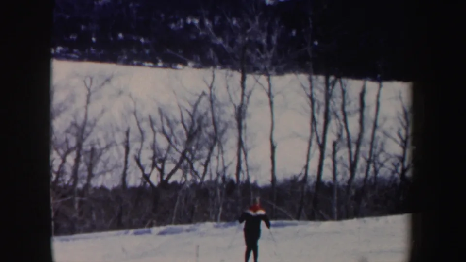 The skier continues downhill across the New Hampshire snowfield, seen small against leafless trees and a dark ridge.