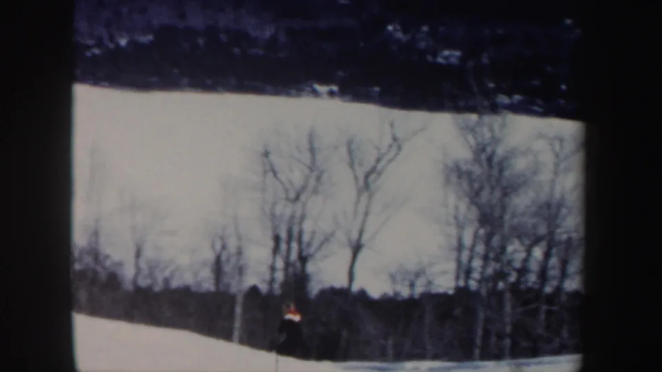 Near the end of the shot, the distant skier remains visible on the snow below a line of bare New Hampshire trees.