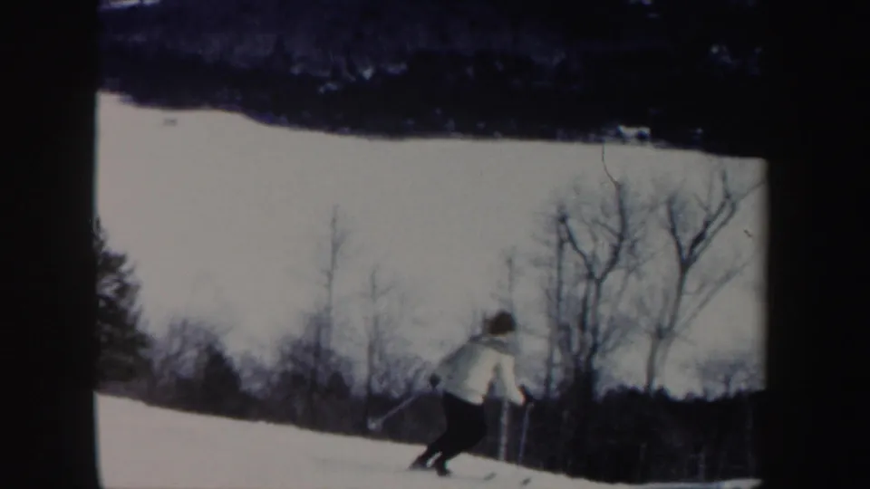 A woman in a white coat skis downhill through a snowy New Hampshire landscape in the 1962 home-movie clip.