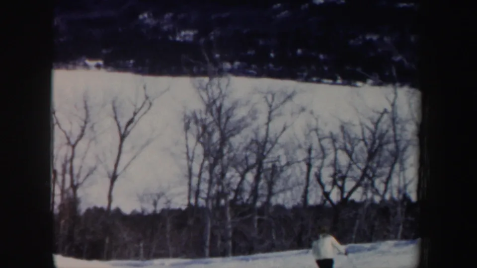 The skier moves low in frame as the handheld 8mm view catches trees and snow on the hillside.