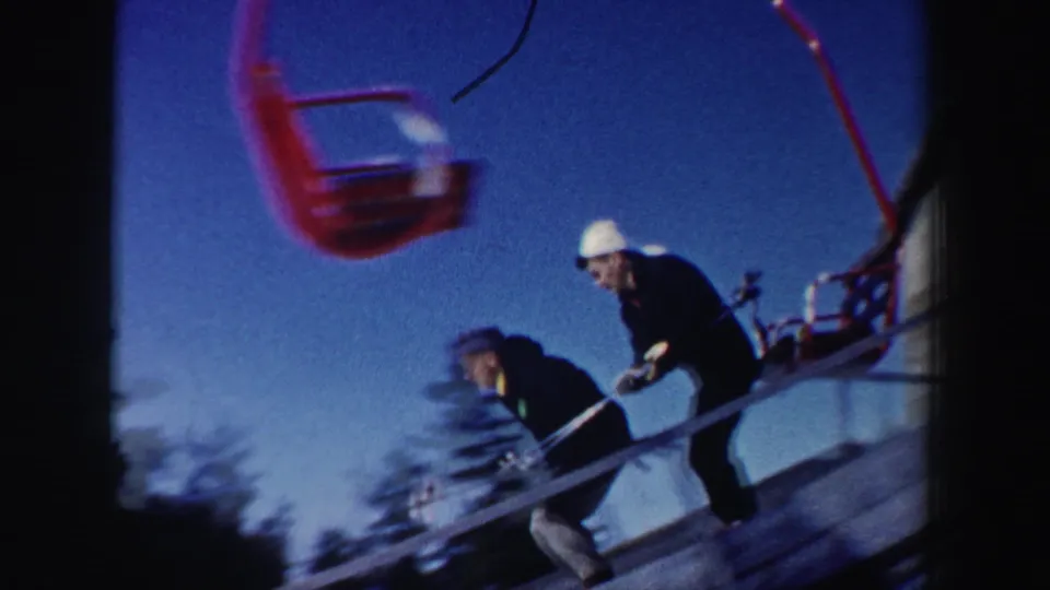 Two skiers near the lift ramp as a red chairlift moves overhead in the New Hampshire ski-area footage.