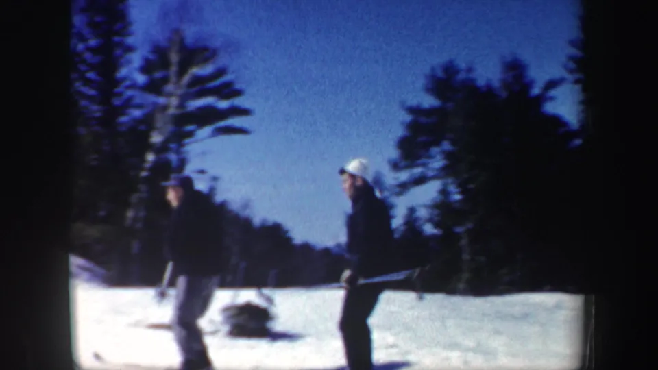 Two skiers with poles stand on a snowy New Hampshire slope with evergreens and winter sky behind them.