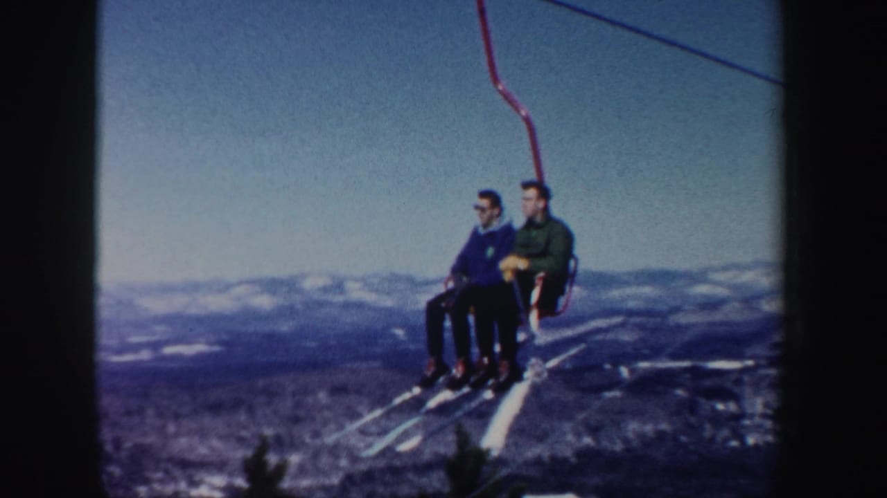 Two men ride a red chairlift with skis hanging over a distant mountain view.