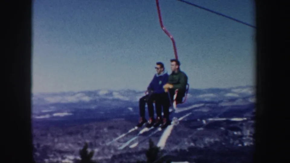 Two adult men ride a red chairlift in New Hampshire, 1962, with long skis dangling above a snowy mountain landscape.