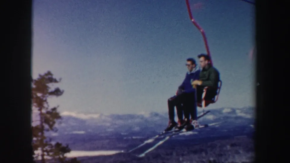 1962 New Hampshire ski footage shows two adult men on a red chairlift against blue sky, snow, mountains, and a tree at frame edge.