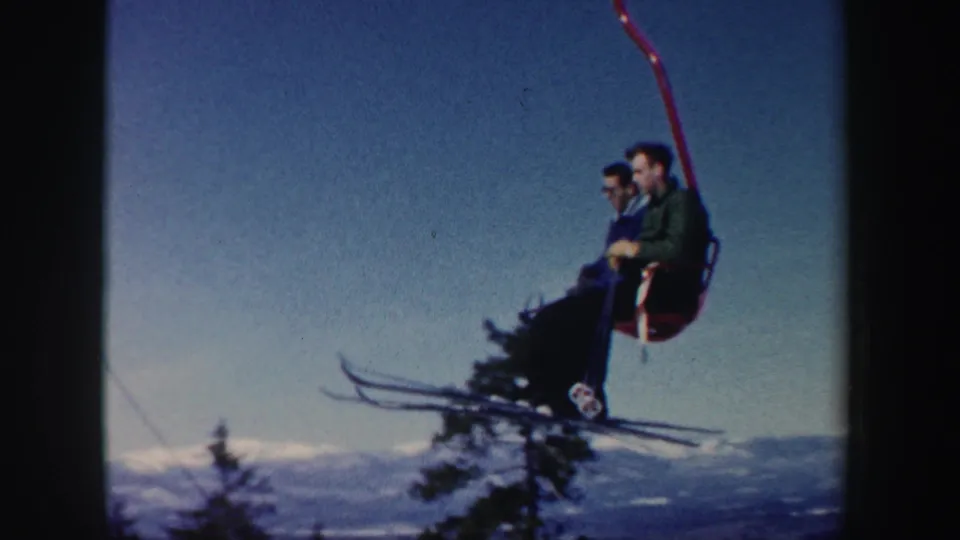 Two adult skiers ride a red chairlift in 1962 New Hampshire, with skis visible against sky, trees, and distant winter hills.