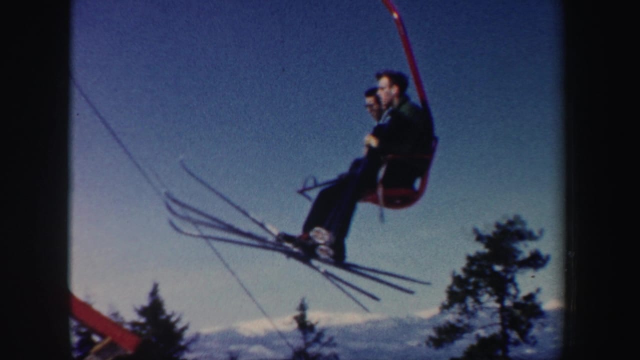 Two skiers ride a red chairlift above trees with skis extending into the blue sky.
