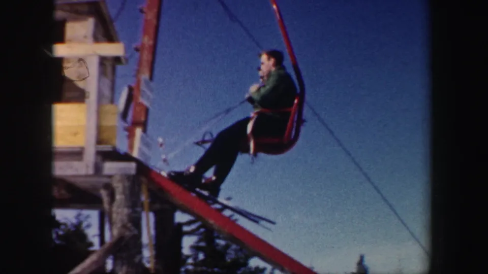 A skier rides a red chairlift toward a wooden landing station in New Hampshire, with lift cables and winter trees visible around the frame.