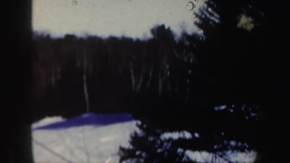 The camera looks down from the lift toward a snowy New Hampshire slope bordered by dark winter trees and soft 8mm frame edges.