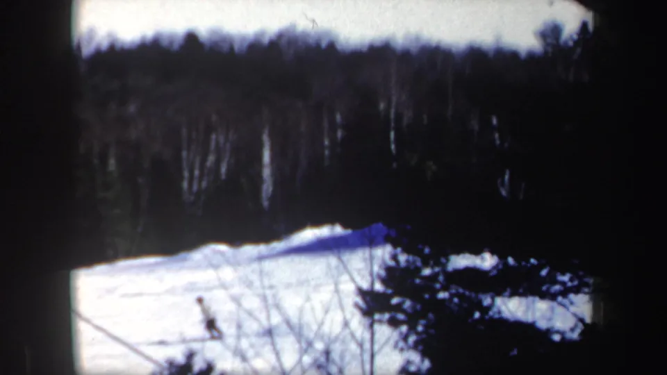 A distant skier crosses a snow-covered slope in 1962 New Hampshire, framed by a dark wooded ridge and visible 8mm film borders.