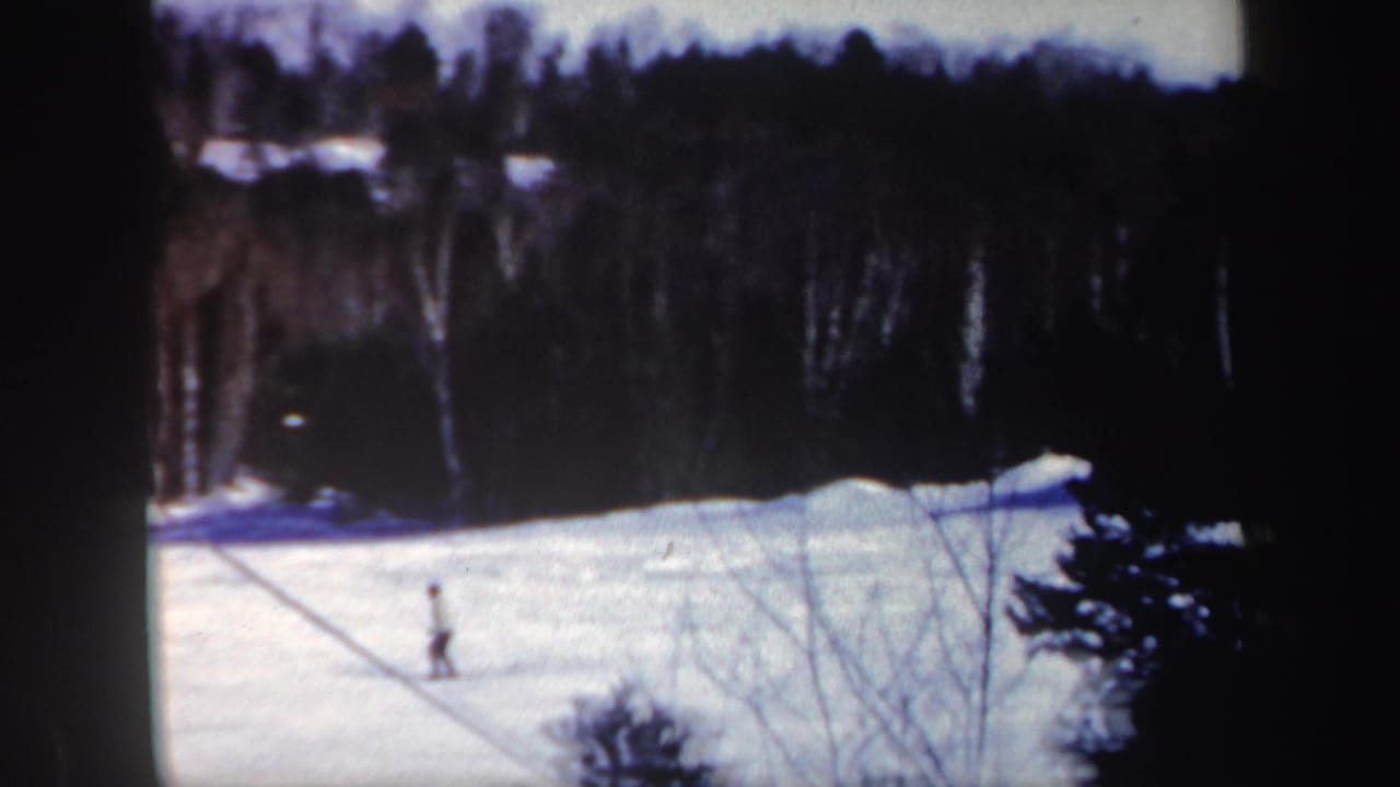 Distant skier crossing a snowy slope bordered by dark trees.