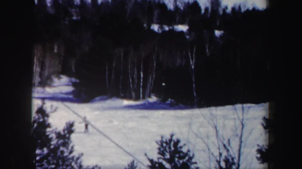 Distant skier on a New Hampshire snow slope in 1962, framed by evergreens and bare woods with soft 8mm color and dark borders.