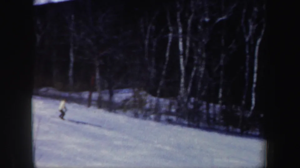 A distant skier crosses a snowy New Hampshire slope in 1962-era 8mm home-movie footage, with dark winter trees behind.