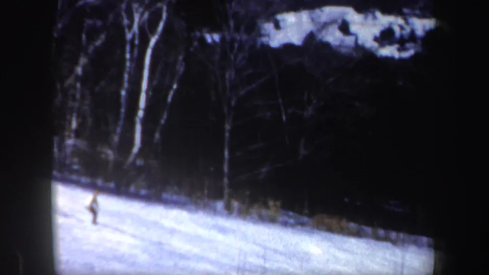 The skier appears small against the bright snow, moving below a dark wooded hillside in New Hampshire home-movie footage.