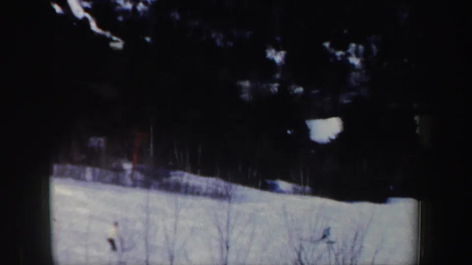 The skier reaches the lower part of the snowy hill, framed by dark trees and visible 8mm edge shadowing.