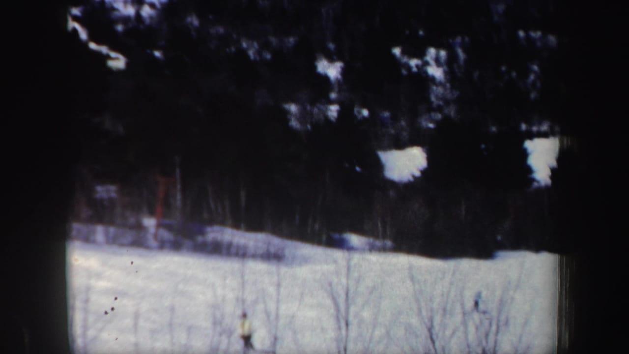 1962 New Hampshire home-movie frame with a small skier on a bright snow-covered slope below a dark tree line.