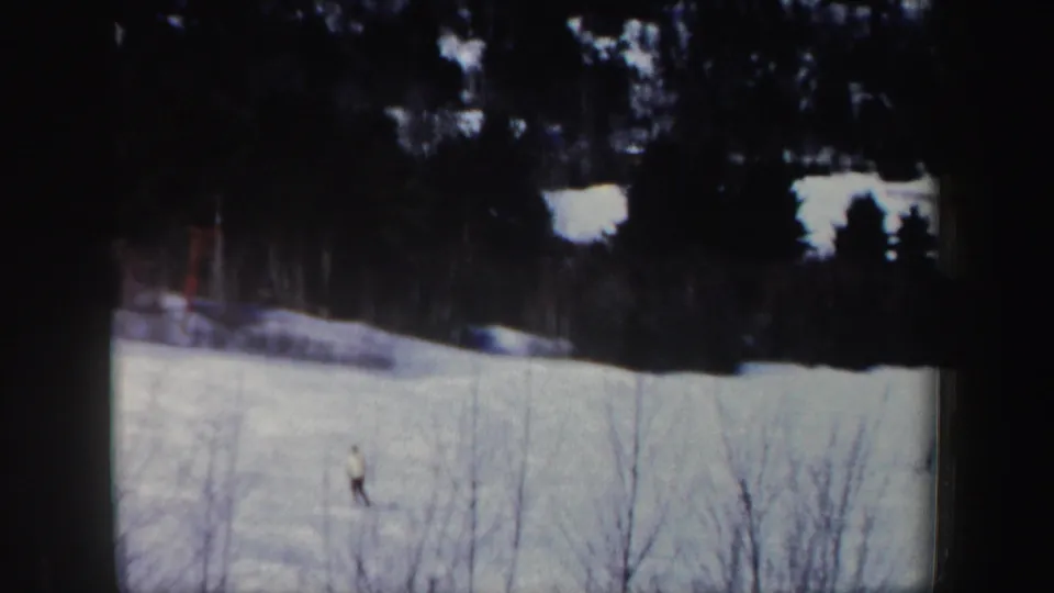 A distant skier moves downhill on a snowy New Hampshire slope, framed by bare trees and dark forest in 1962 home-movie footage.