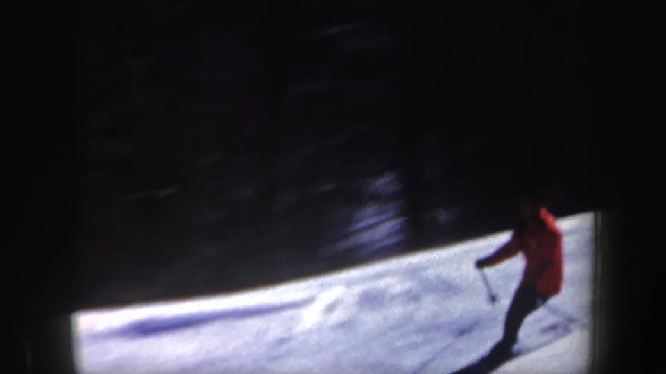 1962 New Hampshire 8mm frame of a skier in red passing along a snowy slope beside a dark wooded area.