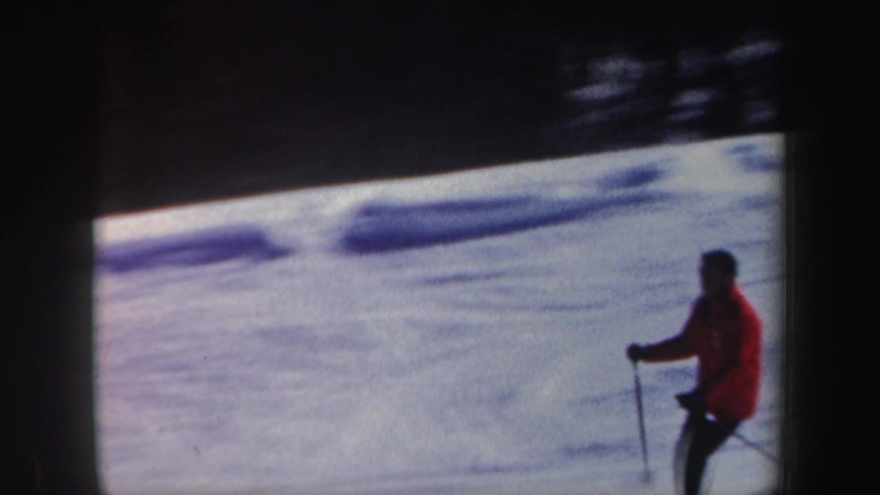In 1962 New Hampshire home-movie footage, an adult skier in a red jacket moves across a snowy slope with poles visible.