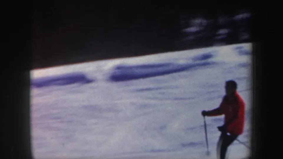 In 1962 New Hampshire home-movie footage, an adult skier in a red jacket moves across a snowy slope with poles visible.