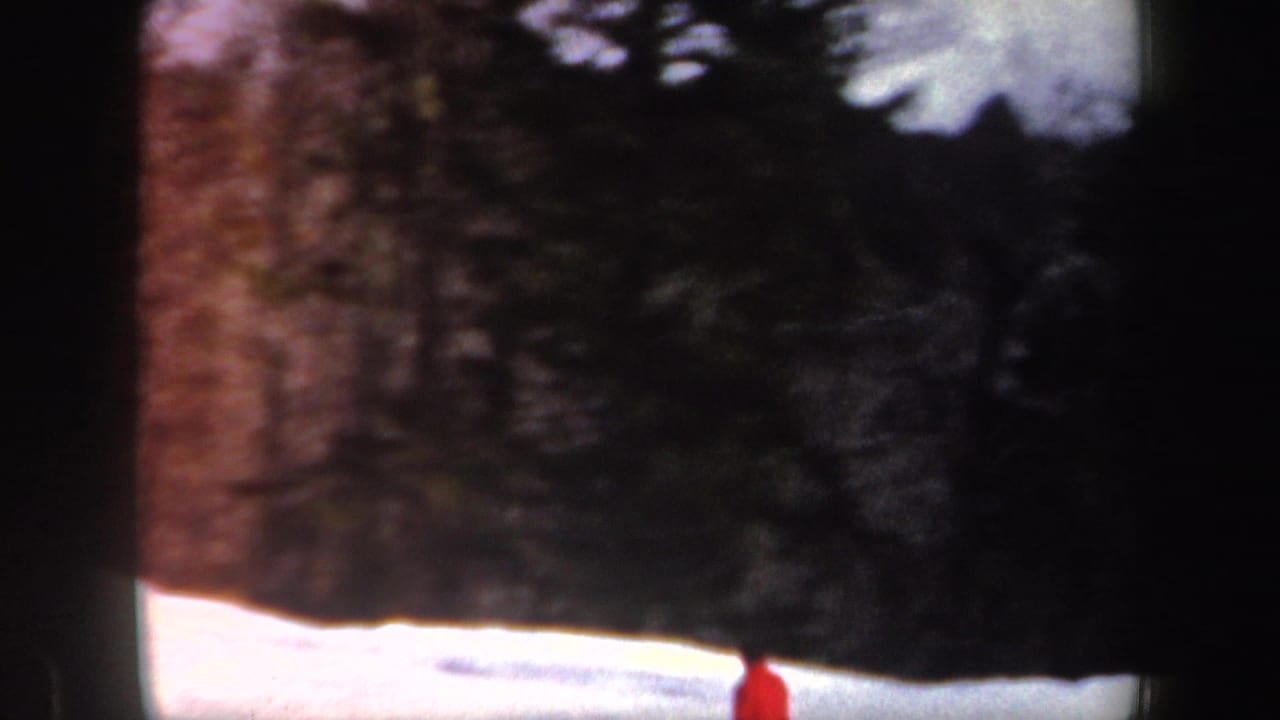 1962 New Hampshire 8mm frame showing a red-coated skier low in the frame beneath a wooded snowy slope.