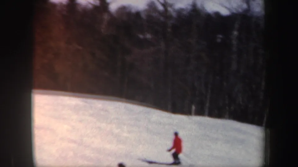 Distant skier in a red coat crosses a sunlit New Hampshire snow slope with dark woods in the background.