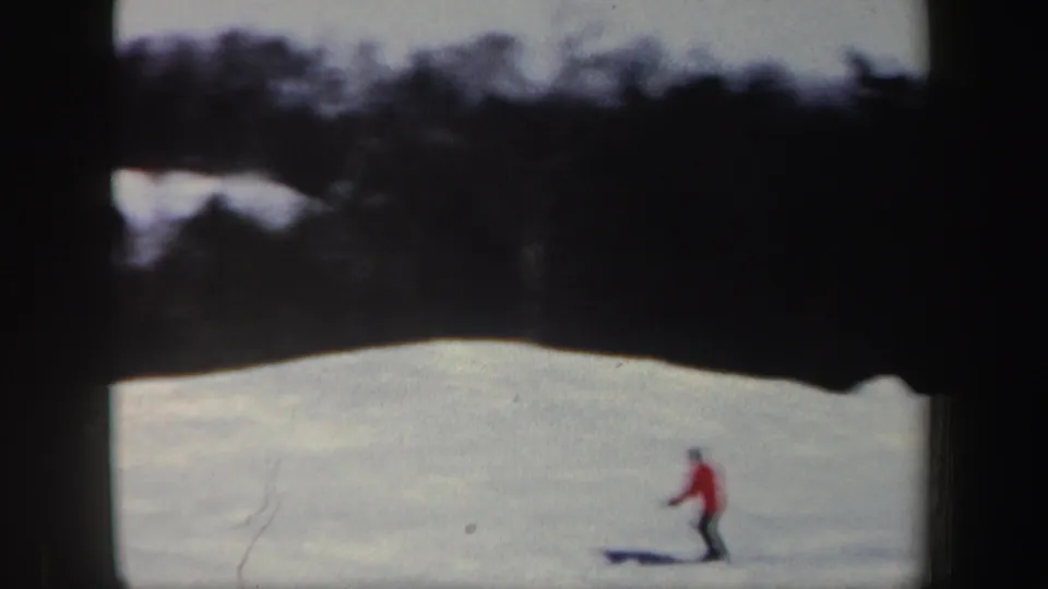 1962 home-movie frame of a red-coated skier on a snowy New Hampshire slope, seen from a distance below woods.