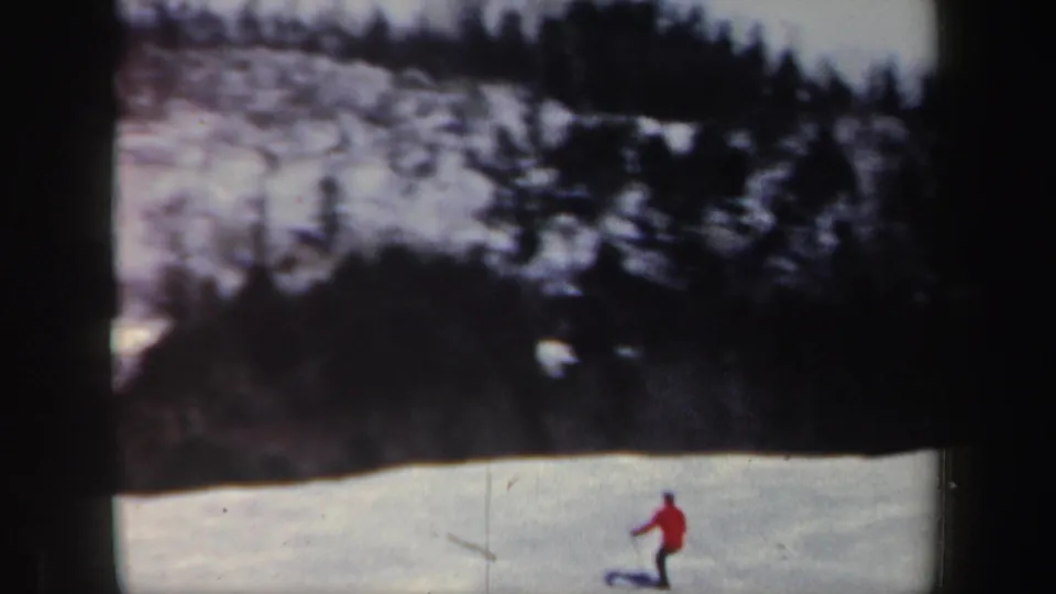 A red-coated skier moves across a snowy New Hampshire slope, framed by wooded hillside and 8mm edge darkness.