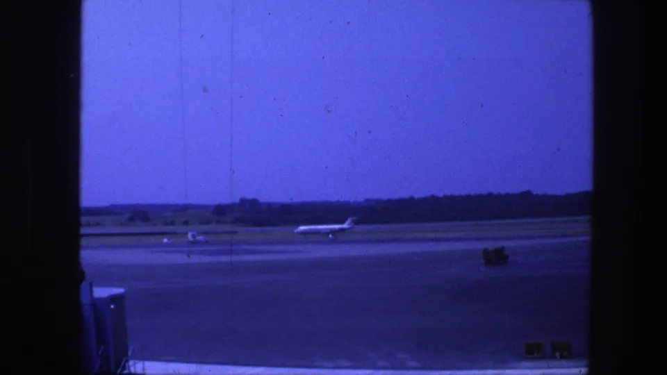 1975 Copenhagen airport home-movie frame showing a high-tail jet taxiing across a runway with trees along the horizon.