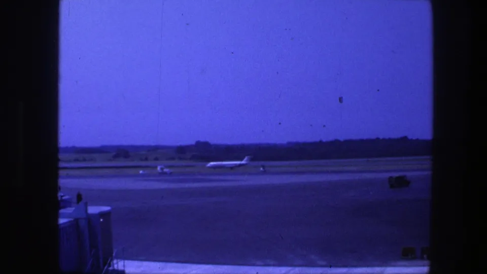 1975 Copenhagen airport frame with a jet taxiing on the runway, a small ground vehicle, and a low tree line beyond the tarmac.