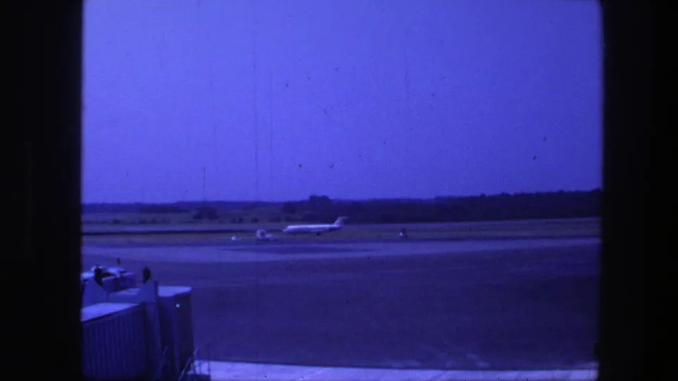 1975 Copenhagen airport home-movie frame showing the jet farther along the runway, with foreground airport equipment and trees beyond.