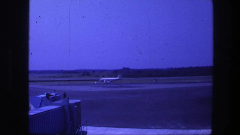 1975 Copenhagen airport frame showing a jet taxiing across the runway, with low airport equipment in front and a tree-lined horizon.