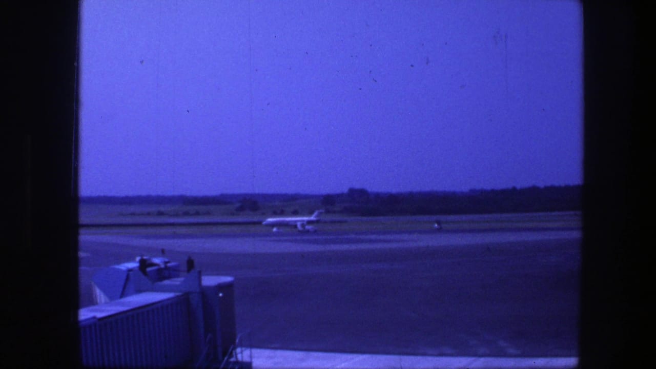 1975 Copenhagen airport view of a commercial jet taxiing beyond a boarding bridge, framed by dark terminal window edges.