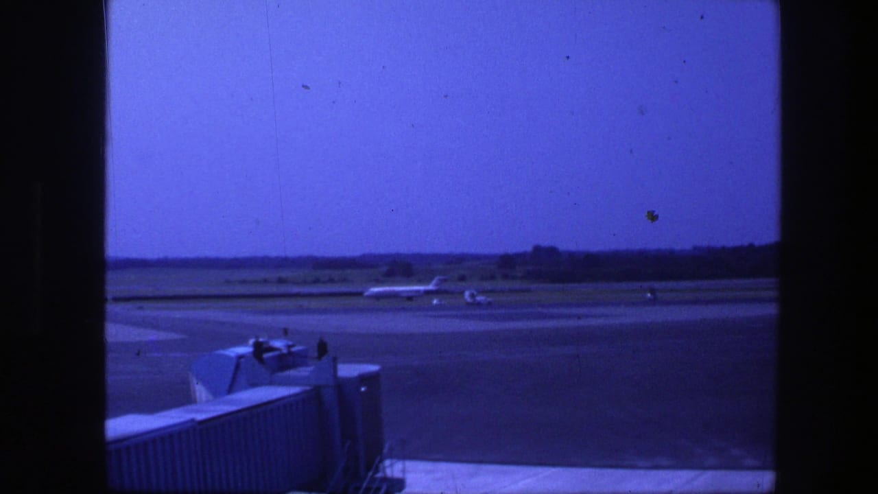 Airliner moving on the runway with terminal window framing and a boarding bridge in the foreground.