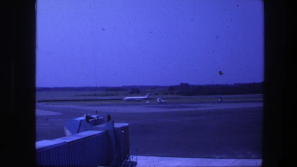 A commercial airliner taxis across the Copenhagen airport runway area under a blue-tinted sky, viewed from inside the terminal.