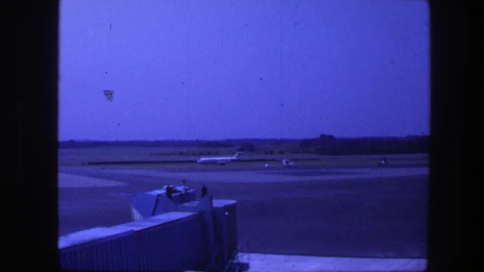 1975 home-movie frame showing a commercial aircraft on the Copenhagen runway area, with terminal structure in the foreground.