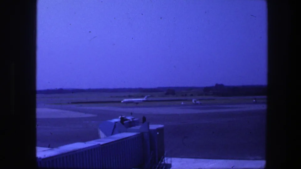 A blue-cast 8mm frame of an airliner taxiing at Copenhagen airport, with the boarding bridge and runway foreground visible.
