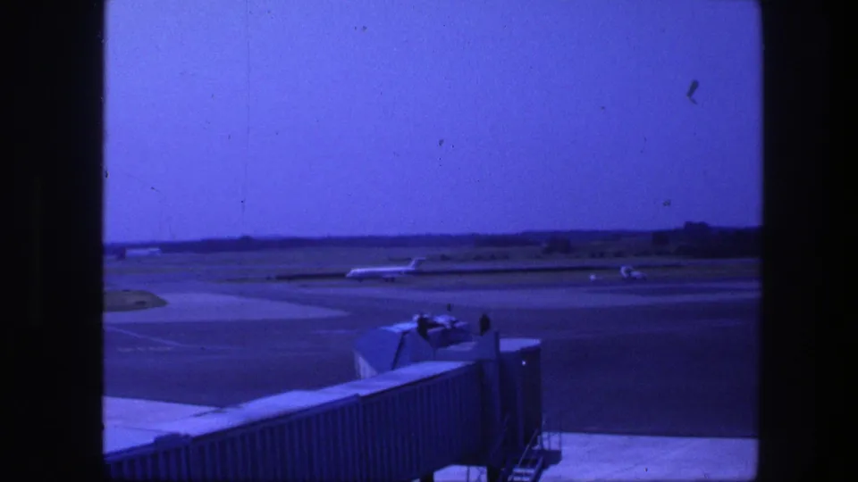 Copenhagen airport tarmac in 1975, viewed past a jet bridge with a distant commercial aircraft on the runway area.