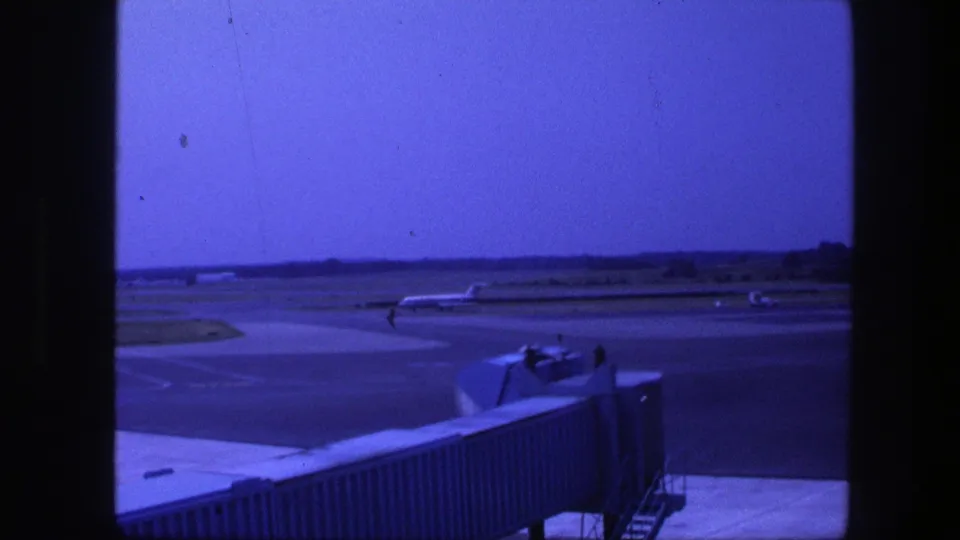 1975 Copenhagen terminal-window footage showing the jet bridge and a distant plane across the blue-tinted airport tarmac.