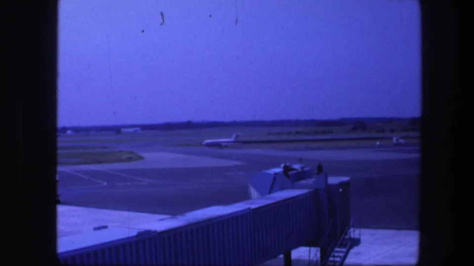 Copenhagen airport in 1975 seen from a terminal window, with a jet bridge foregrounding distant runway activity.