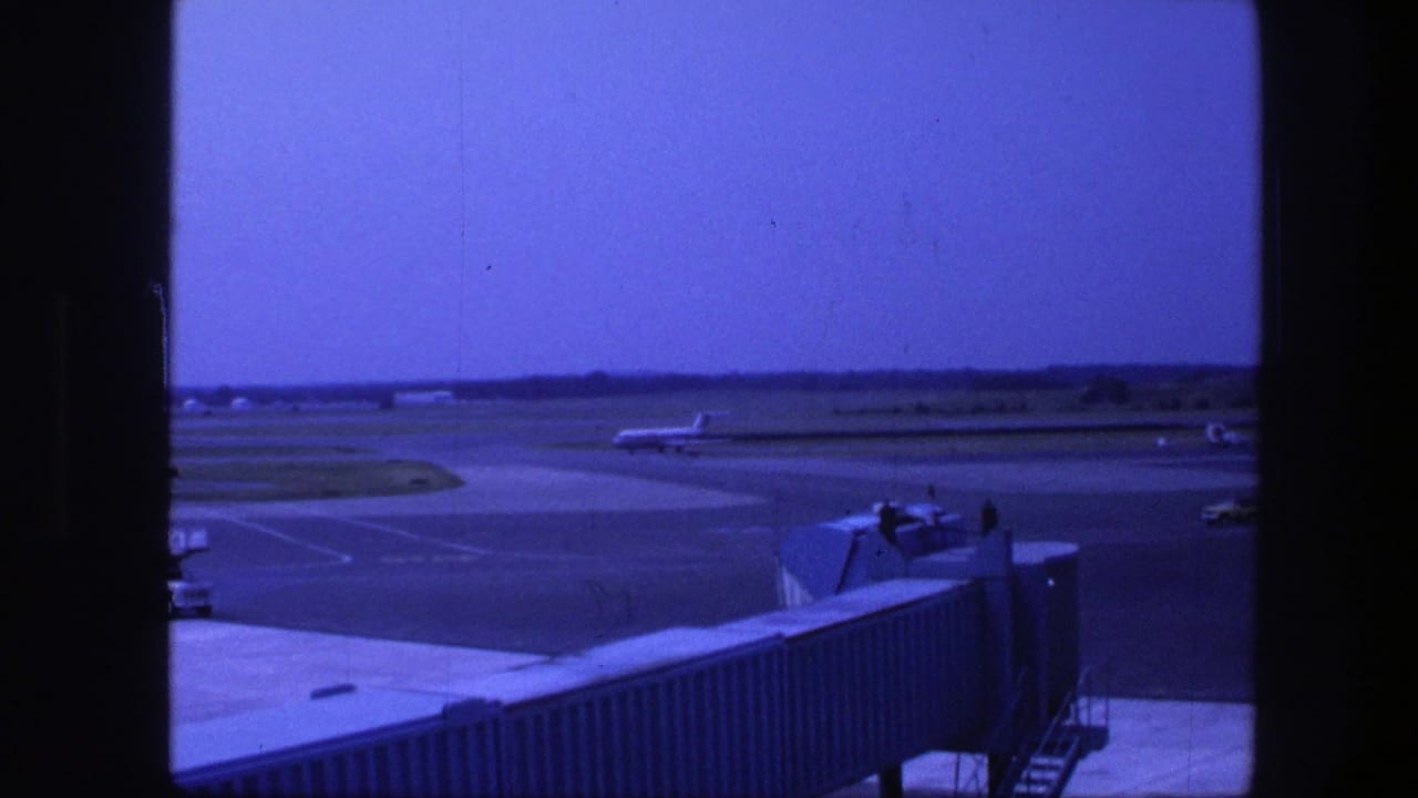 1975 Copenhagen airport view through a terminal window, showing apron pavement, a jet bridge, and distant aircraft shapes.