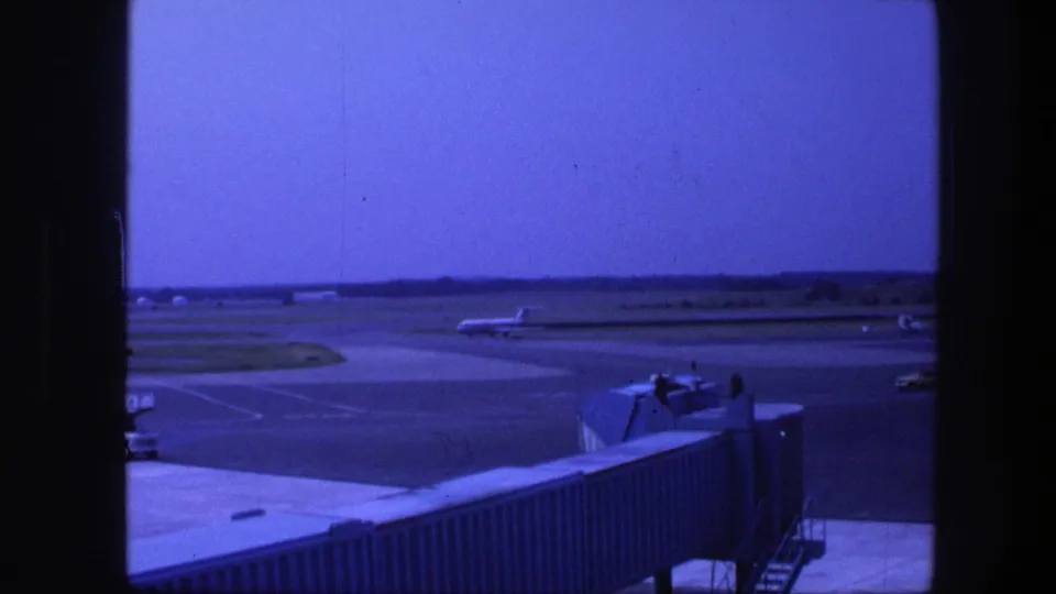 1975 Copenhagen airport view through a terminal window, showing apron pavement, a jet bridge, and distant aircraft shapes.