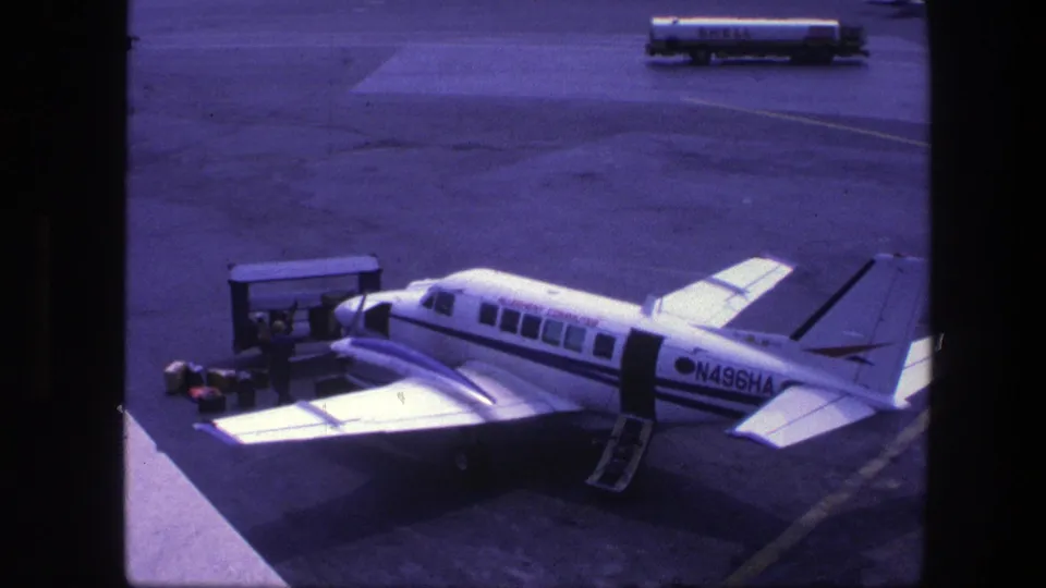 Small white propeller aircraft N496HA at a Copenhagen airport gate in 1975, with stairs down and ground equipment beside it.