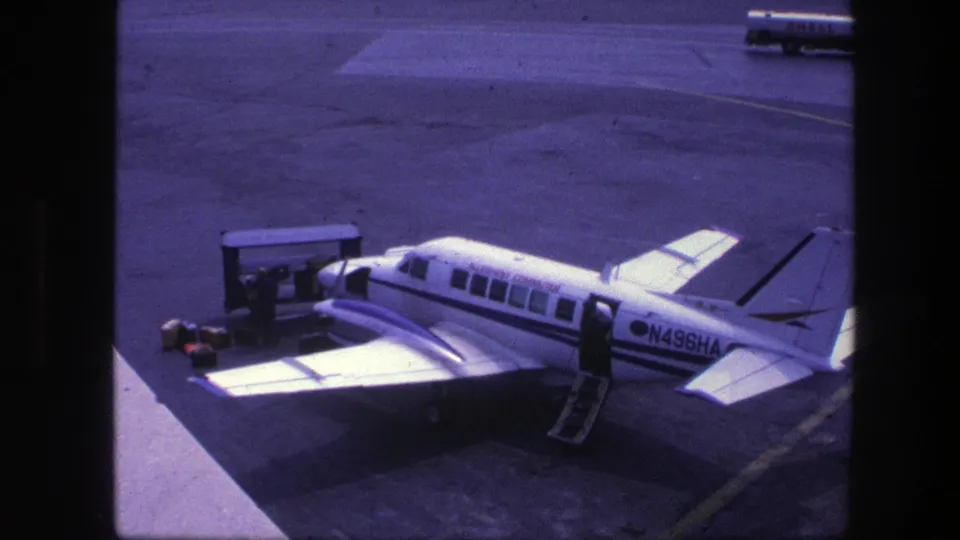 The 1975 Copenhagen airport scene holds on aircraft N496HA, with stairs deployed, ground equipment near the wing, and a Shell tanker beyond.